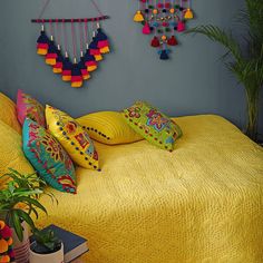 a yellow bed topped with lots of pillows next to a potted plant and wall hangings