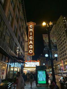 people are walking down the sidewalk in front of some tall buildings at night with lights on