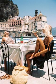 two women sitting at a table on the beach in front of some buildings and water