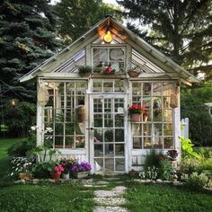 a small white greenhouse with potted plants and flowers in the front window, surrounded by greenery