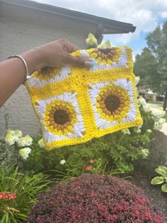 someone holding up a crocheted square with sunflowers on it in front of some flowers