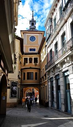 people are walking down an alley way with buildings and a clock tower in the background