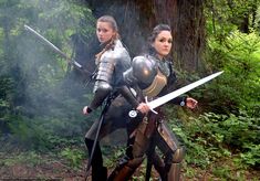 two women dressed in medieval armor holding swords and standing next to each other on a forest path