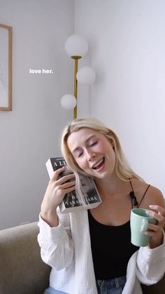 a woman sitting on top of a couch holding a coffee cup and book in her hands