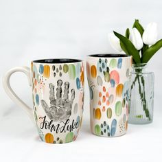 two coffee mugs with hand prints on them and one has a flower in it