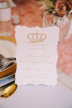 a table topped with a white and gold wedding card next to a glass filled with wine