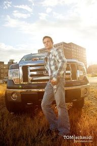 a man leaning on the hood of a truck in an open field with tall buildings behind him