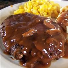 a white plate topped with meat covered in gravy next to potatoes and corn