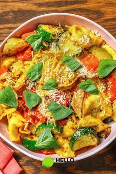 a white bowl filled with pasta and vegetables on top of a wooden table next to a red napkin