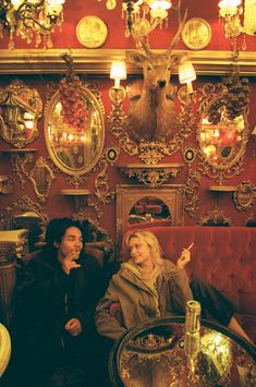 two people sitting on a couch in a room with red walls and gold mirrors above them