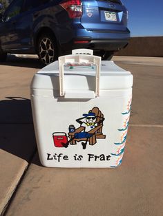 a cooler sitting in the middle of a parking lot next to a blue car with life is frat written on it