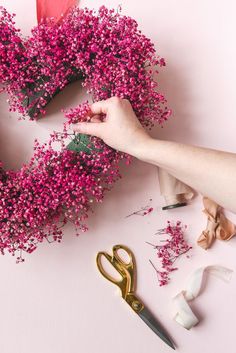 someone is making a heart shaped wreath with pink flowers and scissors on the table next to it