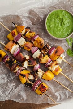 several skewers of meat and vegetables are on a piece of wax paper next to a bowl of pesto