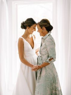 two women standing next to each other in front of a white window holding hands and looking at each other