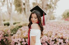 a woman wearing a graduation cap and gown standing in front of pink flowers with red tassels