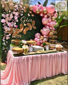 a table topped with lots of pink and gold balloons next to a lush green wall