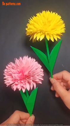 two paper flowers are being held up by someone's hands on a black surface