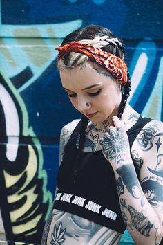 a woman with tattoos standing in front of a graffiti wall holding her hand to her face