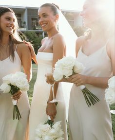 three bridesmaids in white dresses holding bouquets and smiling at each other with the sun shining on them