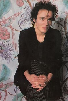 a woman with curly hair sitting in front of a floral wallpapered background wearing a black jacket and striped skirt