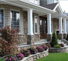 a stone house with white columns and flowers in the front yard on a sunny day