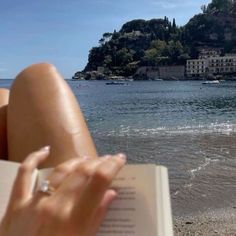 a woman is reading a book on the beach with boats in the water behind her