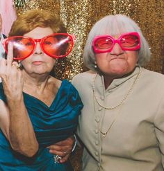 two older women wearing red glasses posing for a photo booth at a party with gold sequins on the wall behind them