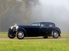 an old black car is parked on the grass in front of some trees and fog