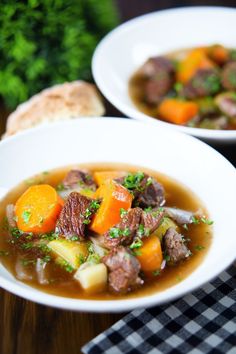 two white bowls filled with stew and carrots on top of a wooden table next to bread