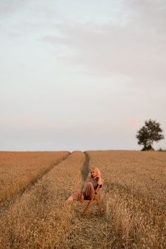 a woman sitting on top of a chair in the middle of a wheat field next to a tree