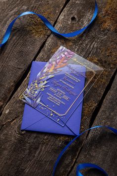 a purple and gold wedding card with blue ribbon on wooden table next to it's envelope