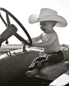 a little boy in a cowboy hat sitting on the steering wheel of a tractor