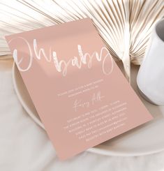 a pink and white wedding card on top of a plate next to a vase with palm leaves