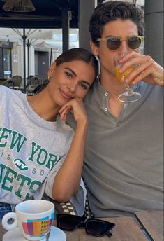 a man and woman sitting at a table with drinks in front of them, both holding their arms around each other