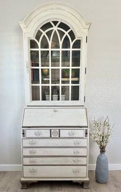 a white dresser with glass doors on top and drawers below it next to a blue vase filled with flowers