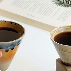 two cups of coffee sitting on top of a table next to an open book and a feather