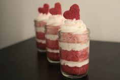 three red velvet desserts with white frosting and heart shaped decorations in jars on a table