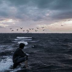 a man kneeling in the water watching birds fly overhead on a gloomy day at sea