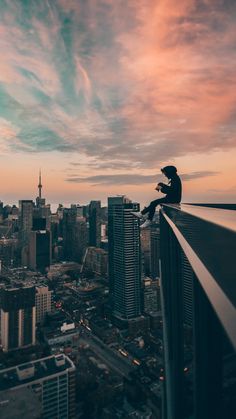 a man sitting on top of a tall building next to a city skyline at sunset