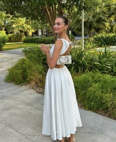 a woman in a white dress is standing on the sidewalk with her hands together and looking at the camera