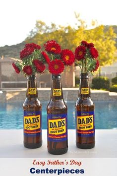 three beer bottles with red flowers in them sitting on a table next to a pool