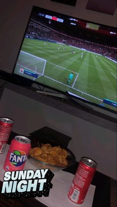 a tv that has some food on the table in front of it and two cans of sodas next to it