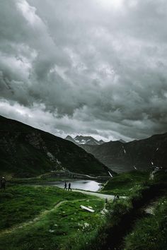 some people are standing in the grass near a body of water with mountains in the background