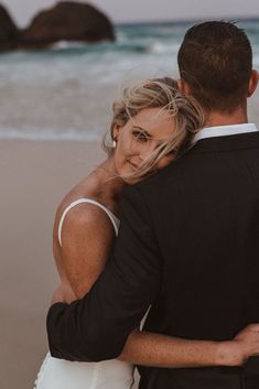 a bride and groom hugging on the beach in front of an ocean with large rocks