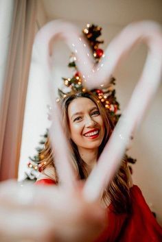 a woman is smiling and making a heart shape with her hands while standing in front of a christmas tree