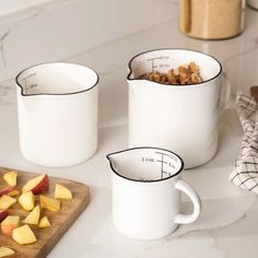 three white mugs filled with fruit on top of a counter next to a cutting board