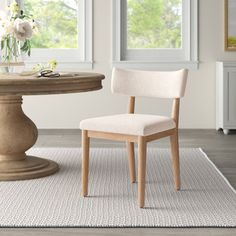 a white chair sitting next to a table on top of a rug in front of two windows