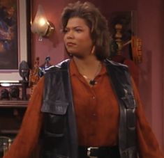 a woman standing in a living room wearing a brown shirt and black leather vest with her hands on her hips