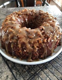 a chocolate bundt cake with icing and nuts on a white plate sitting on a cooling rack