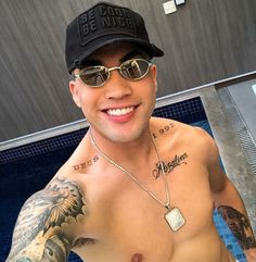 a shirtless man wearing sunglasses and a hat with tattoos on his chest stands in front of a swimming pool